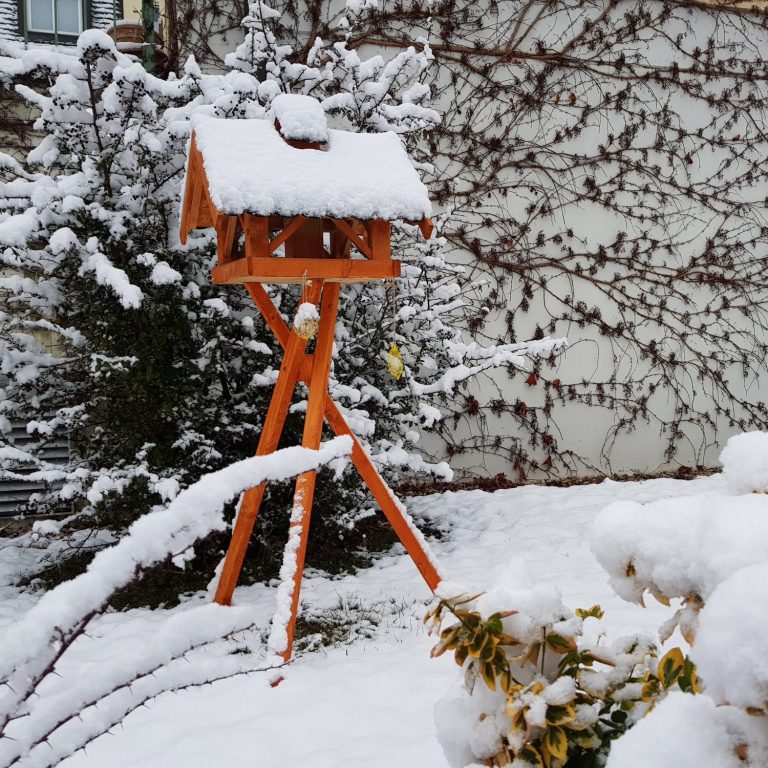 Vogelhaus mit prakitischem Dreibeinständer, umgeben von Schnee und winterlicher Landschaft.